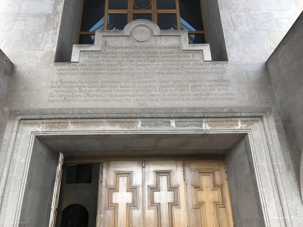 Große hölzerne Tür vor einem steinernen Gebäude mit einem Glasfenster darüber, auf dem likely eine Inschrift für ein Holocaust-Mahnmal zu sehen ist.