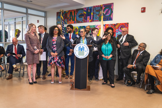 Gruppe von Menschen vor einem Podium mit einem Mikrofon und einer Tafel mit dem Text "Sancho City Councilman Joins City Council", mit sitzenden und stehenden Teilnehmern, Flaggen, einem Feuerlöscher und Deckenleuchten im Hintergrund.