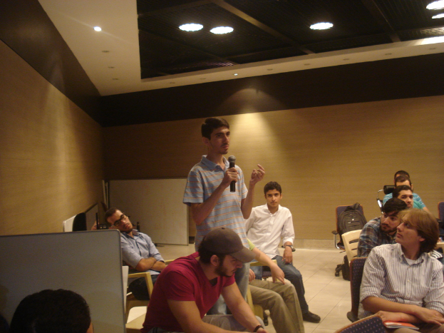 Eine Person steht in einem Büroraum und hält ein Mikrofon, während mehrere andere auf Stühlen sitzen und zuhören.