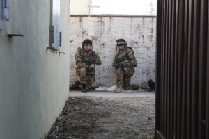 Eine Szene mit einer Wand und einem Fenster auf der linken Seite, einer Eisenwand auf der rechten Seite, zwei Soldaten und Erde in der Mitte und Gebäuden, einem Strommast und einer weiteren Wand im Hintergrund.