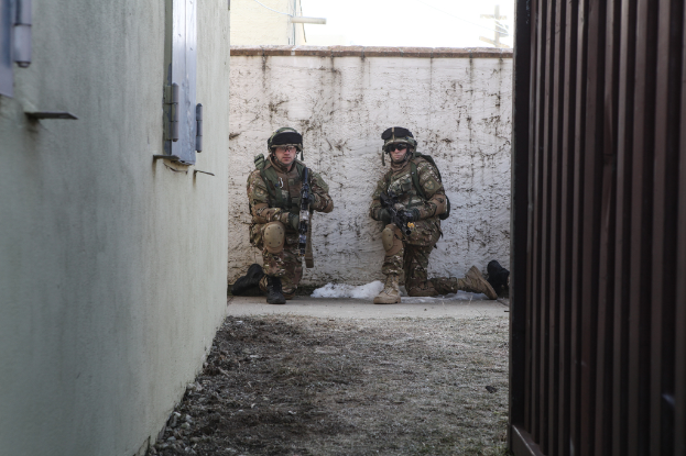 Eine Szene mit einer Wand und einem Fenster auf der linken Seite, einer Eisenwand auf der rechten Seite, zwei Soldaten und Erde in der Mitte und Gebäuden, einem Strommast und einer weiteren Wand im Hintergrund.