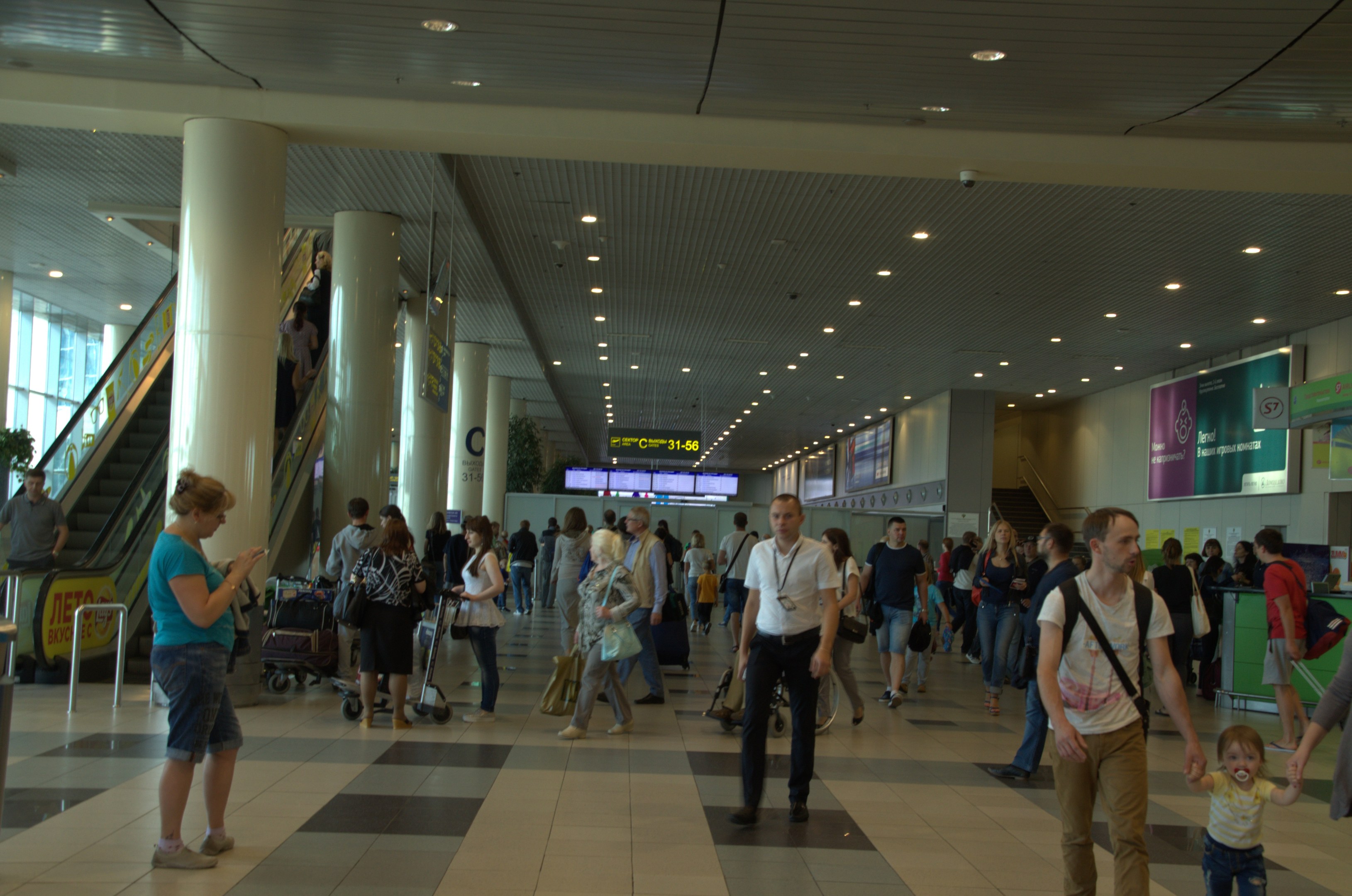 Eine große Gruppe von Menschen, die durch einen Flughafen gehen, einige tragen Gepäck und Kofferwagen, mit Rolltreppen, Säulen, Texttafeln, Deckenleuchten und Glaswänden im Hintergrund.