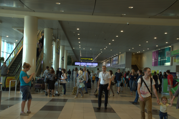 Eine große Gruppe von Menschen, die durch einen Flughafen gehen, einige tragen Gepäck und Kofferwagen, mit Rolltreppen, Säulen, Texttafeln, Deckenleuchten und Glaswänden im Hintergrund.