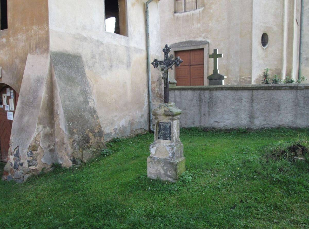 Alte Kirche mit einem Kreuz davor, umgeben von Gras und Pflanzen, mit Fenstern, einer Tür und Papieren an der Wand.