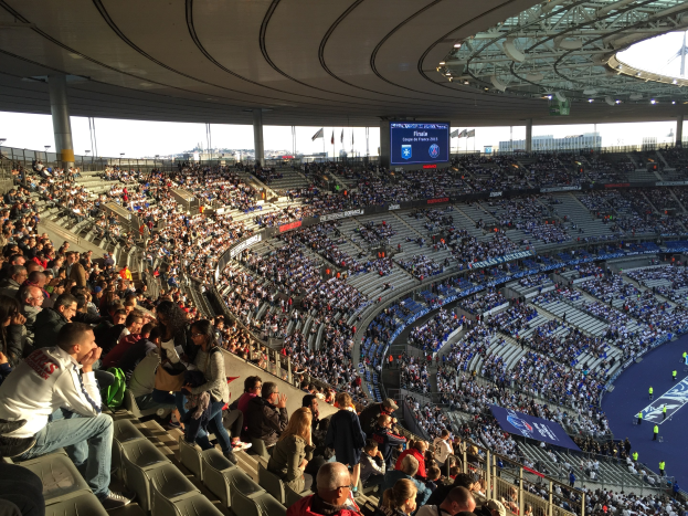 Große Zuschauermenge im Allianz Arena Stadion bei einem Fußballspiel, mit einer Bühne auf der rechten Seite, Fahnen, Stangen, einem Bildschirm und sichtbarem Himmel im Hintergrund.