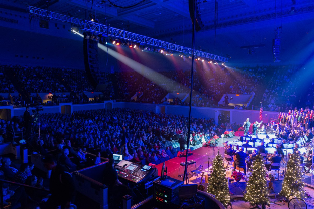 Eine große Menschenmenge in einem geschmückten Auditorium schaut einer Band auf einer Bühne mit Weihnachtsbäumen, Lautsprechern und festlicher Beleuchtung zu.