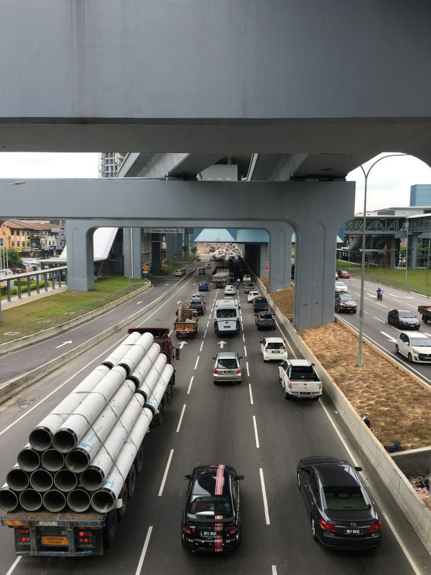 Eine stark befahrene Autobahn mit Schwerlastverkehr unter einer Brücke, umgeben von Gras, Bäumen, Gebäuden, Masten, Laternen und einem klaren Himmel.