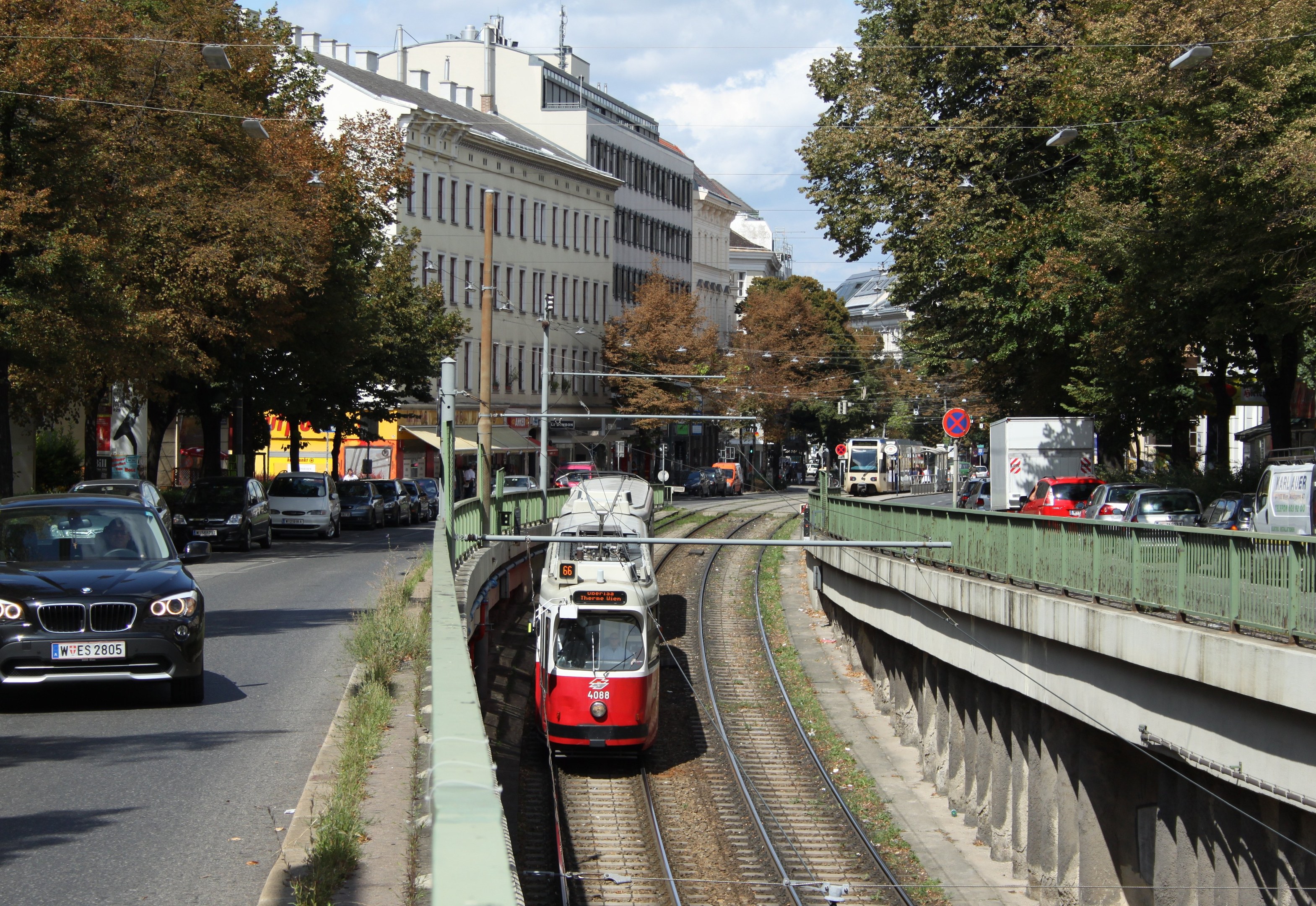 Rote und weiße Straßenbahn auf Schienen neben einer Stadtstraße mit Fahrzeugen, Gebäuden, Bäumen und Masten im Hintergrund.