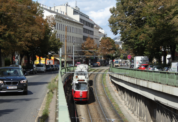 Rote und weiße Straßenbahn auf Schienen neben einer Stadtstraße mit Fahrzeugen, Gebäuden, Bäumen und Masten im Hintergrund.