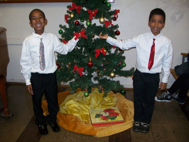 Ein geschmückter Weihnachtsbaum steht in einem Raum mit einem Holztisch links und einem Stuhl rechts, zwei Jungen in weißen Hemden lächeln und halten den Baum, eine Kiste und andere Gegenstände liegen auf dem Boden, und eine weiße Wand mit angebrachten Gegenständen im Hintergrund.