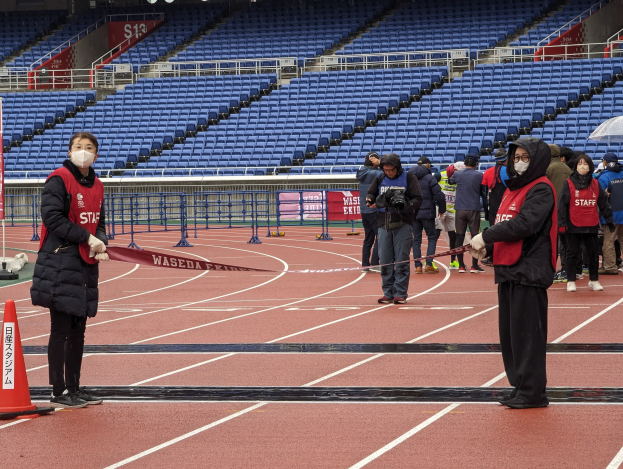 Gruppe von Menschen, die auf einer Bahn stehen und ein Band halten, mit einem Verkehrskegel links und eventbezogenen Gegenständen im Hintergrund.