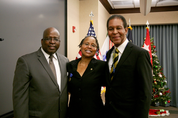 Eine Gruppe strahlender Menschen vor einem geschmückten Weihnachtsbaum mit verschiedenen Gegenständen, Fahnen und einem Vorhang im Hintergrund, mit einem Tuch und zusätzlichen Gegenständen auf der rechten Seite.