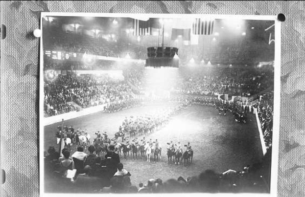 Ein Schwarz-Weiß-Foto einer großen Menschenmenge in einem Stadion, mit einigen Menschen auf Pferden und anderen Stehenden, einer Bühne mit Fahnen und Lichtern in der Mitte und Text unten.