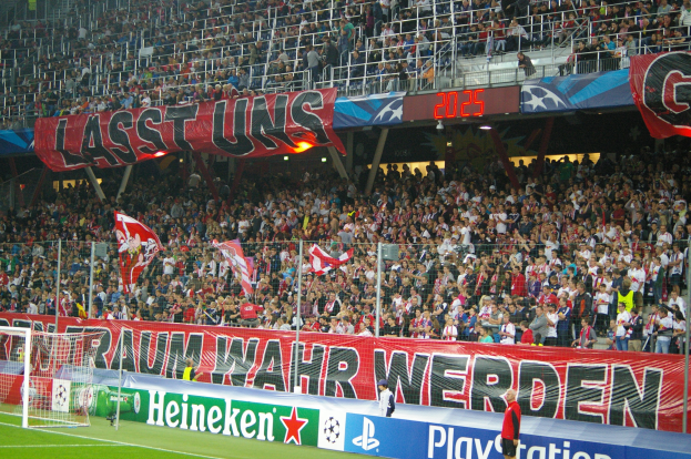 Fußballspiel in einem Stadion mit großer Menschenmenge, saftig grünem Rasen, Torpfosten links, Bodenbanner, Menschen mit Fahnen und Hintergrundgeländer, Tafeln, Lampen und andere Gegenstände.