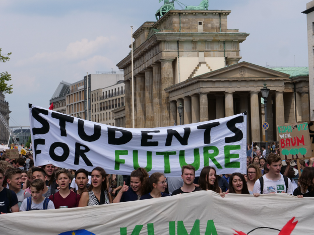 Gruppe von Schülern marschiert in Berlin mit einer leuchtend bunten "Students for Future"-Fahne vor einem Hintergrund aus Gebäuden, Bäumen und Himmel.