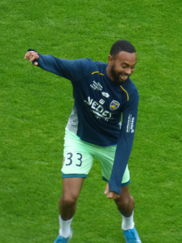 Ein Mann in einem blauen Hemd und grünen Shorts steht auf einem Fußballfeld, lächelt und trägt Schuhe, bereit zu spielen.