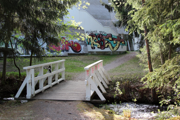 Holzbrücke über einen Bach vor einem Graffiti-bedeckten Gebäude, umgeben von grünem Gras und Bäumen mit fließendem Wasser darunter.