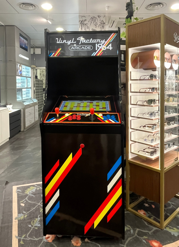 Ein Geschäft mit einem vintage Arcade-Automaten in der Mitte, einem Glasvitrinenschrank auf der rechten Seite und Retro-Stil-Beleuchtung mit Schränken und einem Fernseher im Hintergrund.