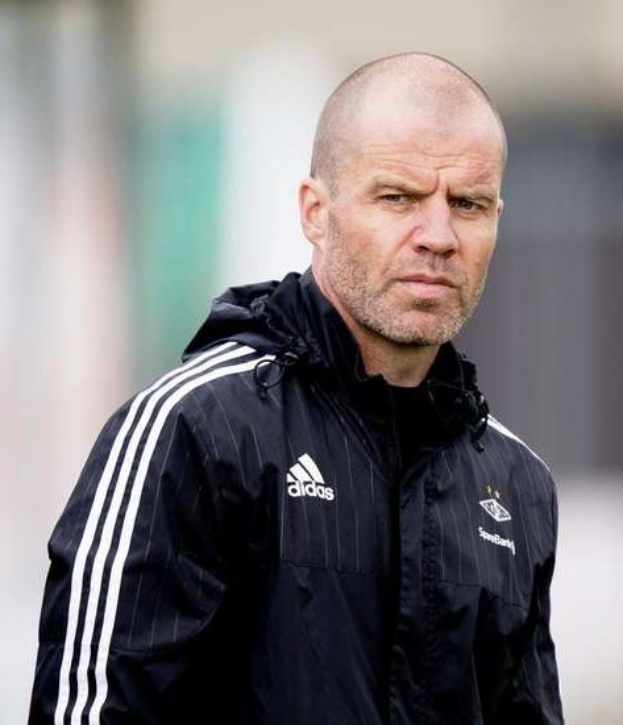 Bald coach in einem blauen Jackett mit weißen Streifen und weißem Kragen, der auf einem Fußballfeld steht und die Hände zusammenlegt.