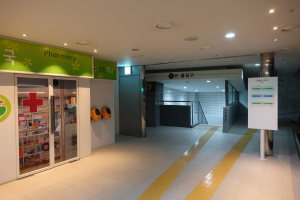 Eingang einer Apotheke mit einem "Apotheke"-Schild auf der linken Seite, einer Tafel mit Text auf der rechten Seite und beleuchteten Lampen über dem Dach.