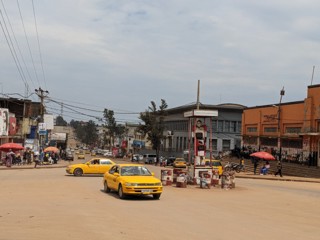 Ein gelbes Taxi fährt eine belebte Stadtstraße mit Gebäuden an den Seiten hinunter, während Fußgänger mit Regenschirmen auf dem Bürgersteig gehen, unter einem bewölkten Himmel, mit Strommasten und Drähten im Hintergrund.