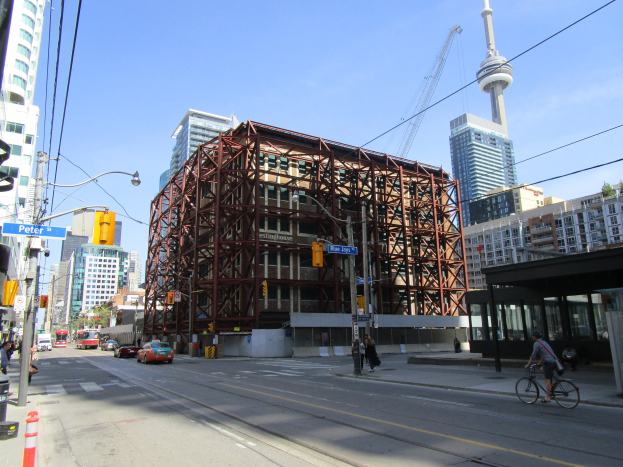 Eine belebte Stadtstraße mit einem großen Gebäude im Hintergrund, das unter Bauarbeiten steht, mit Fahrzeugen, einem Radfahrer, Fußgängern, Ampeln, Pfählen, Schildern, einem Kran und Gerüsten.