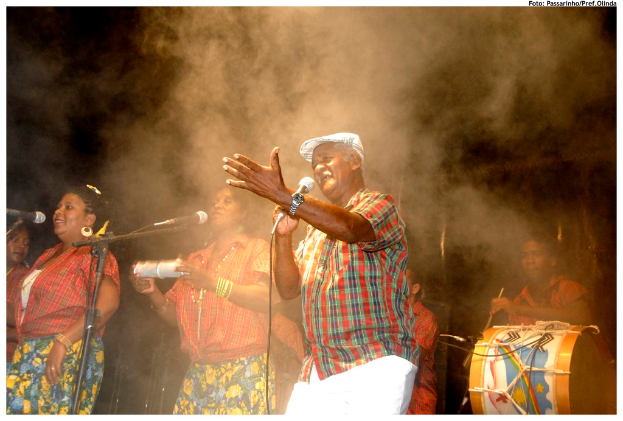 Eine Gruppe von Musikern, die auf der B├╝hne stehen, mit einem Mann, der in die Mitte singt und einem anderen Mann, der rechts ein Schlagzeug spielt.