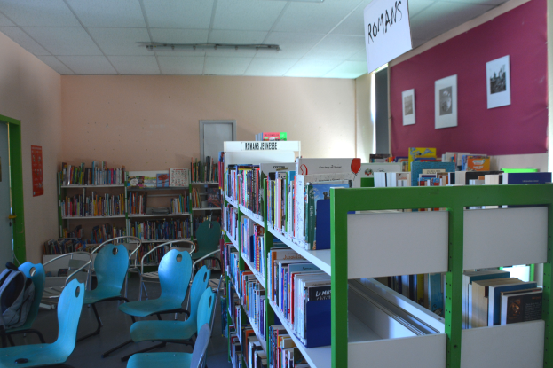 Ein Bibliotheksinneres mit Bücherregalen, Stühlen, Taschen auf dem Boden, Wandrahmen, einer Texttafel, einer Tür und Deckenleuchten.