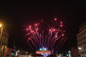 Eine belebte Stadtstraße bei Nacht mit Menschen, Fahrzeugen, Zelten und Gebäuden sowie einem farbenfrohen Feuerwerk am Himmel.