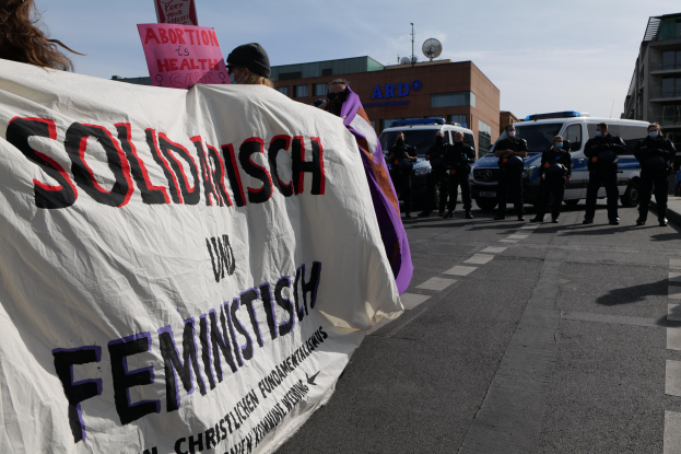 Eine Gruppe von Menschen marschiert auf einer Straße und hält ein Banner mit der Aufschrift "Solidarität und Feminismus", mit parkenden Fahrzeugen und Gebäuden im Hintergrund unter einem klaren blauen Himmel.