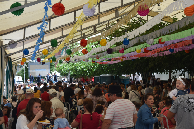 Eine große Menschenmenge sitzt an Tischen unter einem Zelt, einige tragen Mützen und andere halten Teller mit Essen, mit Bäumen im Hintergrund und dekorativen Gegenständen, die von der Decke hängen.