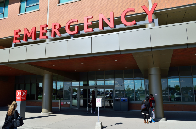 Eine Gruppe von Menschen geht vor einem Glaswand-Notfallgebäude mit Säulen, einem Namensschild, Schildern an Pfosten und einem Mülleimer vorbei.