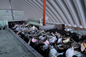 Großer Haufen Müll auf einem Förderband in einer Recyclinganlage mit einer Maschine und einem Metalldach im Hintergrund.