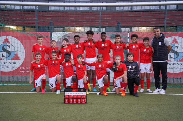 Eine Gruppe junger Männer in roten T-Shirts und schwarzen Shorts steht auf einem Fußballfeld neben einer Kiste roter Gegenstände, mit Bannern im Hintergrund, die "FC Rot-Schwarz Eglaute - Champions League" lesen.