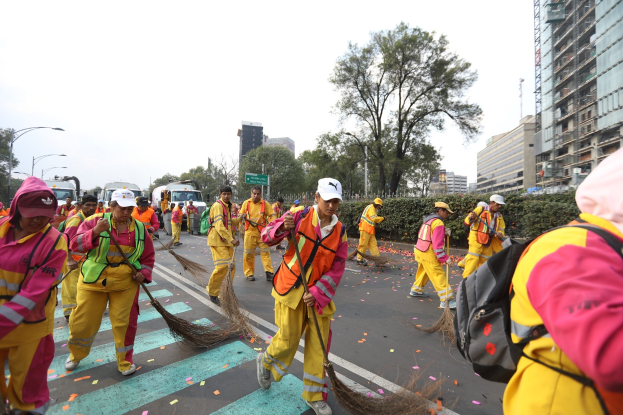Eine Gruppe von Menschen in gelben Uniformen, Mützen und mit Taschen, die die Straße mit Besen kehren, während Fahrzeuge vorbeifahren, mit Bäumen, Gebäuden, Laternenpfählen und einem klaren blauen Himmel im Hintergrund.