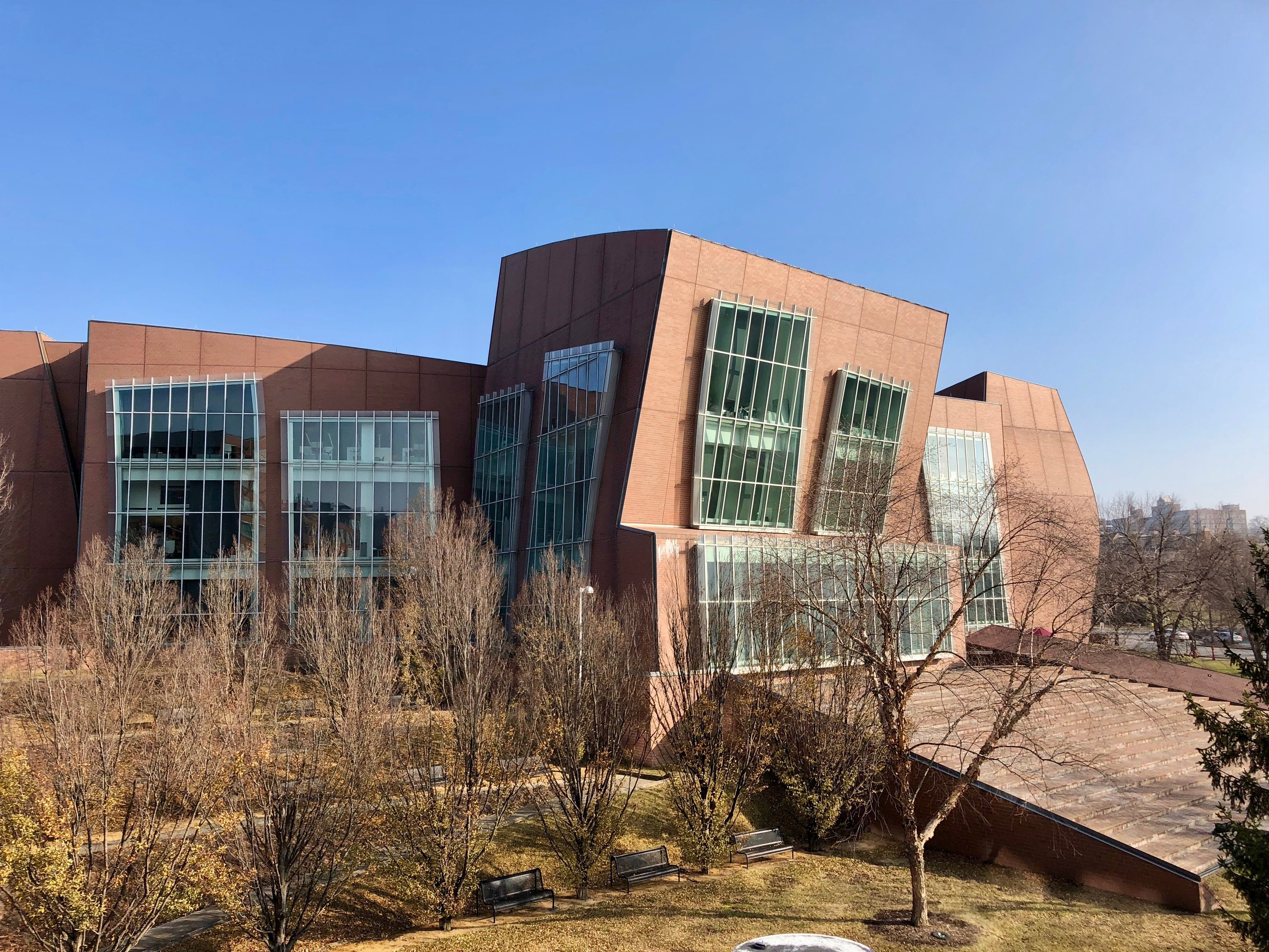 Großes Gebäude mit vielen Fenstern, umgeben von Bäumen, Bänken, Gras und einem Lichtmast, mit anderen Gebäuden und einem klaren blauen Himmel im Hintergrund, als der neue Campus der University of Illinois at Chicago identifiziert.
