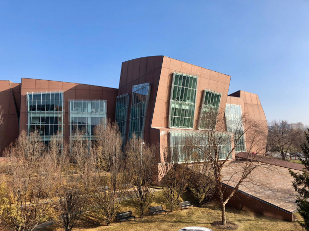 Großes Gebäude mit vielen Fenstern, umgeben von Bäumen, Bänken, Gras und einem Lichtmast, mit anderen Gebäuden und einem klaren blauen Himmel im Hintergrund, als der neue Campus der University of Illinois at Chicago identifiziert.