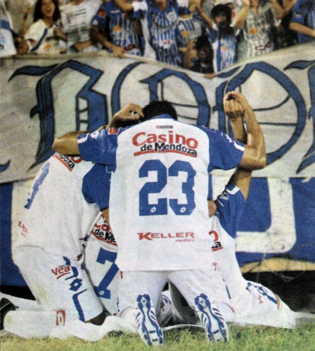 Eine Gruppe von Fußballspielern feiert ein Tor auf dem Boden vor einer jubelnden Menge, mit einem Banner im Hintergrund.