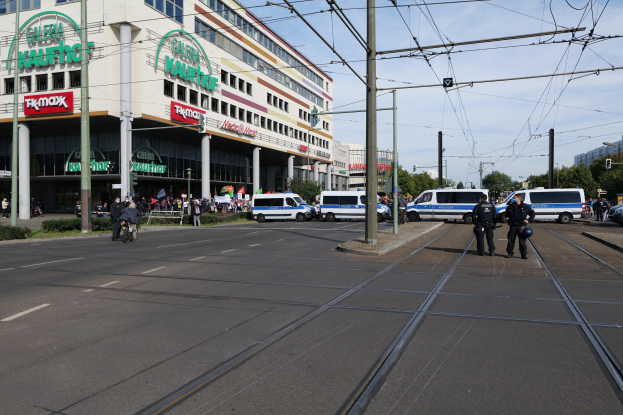 Polizisten stehen neben einer Straßenbahn auf der Straße mit Autos, Strommasten, Gebäuden, Bäumen und einem bewölkten Himmel im Hintergrund.