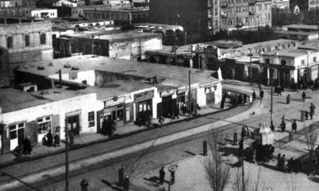 Schwarzes und weißes Foto einer Stadtstraße mit Fußgängern, Gebäuden, Bäumen, Strommasten und Drähten, die das historische Stadtbild widerspiegeln.