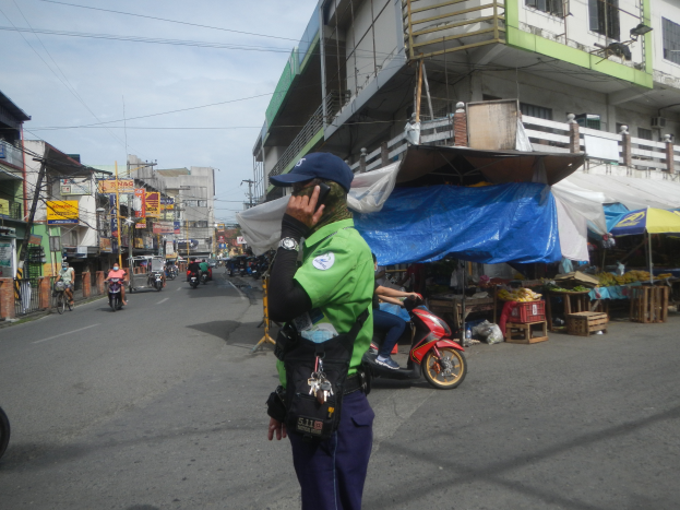 Ein Polizeibeamter in grüner und blauer Uniform und blauer Mütze steht an der Straßenseite und spricht auf einem Handy, mit fahrenden Fahrzeugen im Hintergrund, Ständen mit Gegenständen rechts und einem bewölktem Himmel.