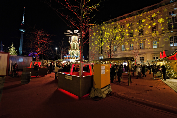 Ein belebter Weihnachtsmarkt in Berlin, Deutschland mit Menschen um dekorierte Stände, festliche Lichter, Bäume, Gebäude, Laternenmasten und einen Turm unter einem dunklen Himmel.