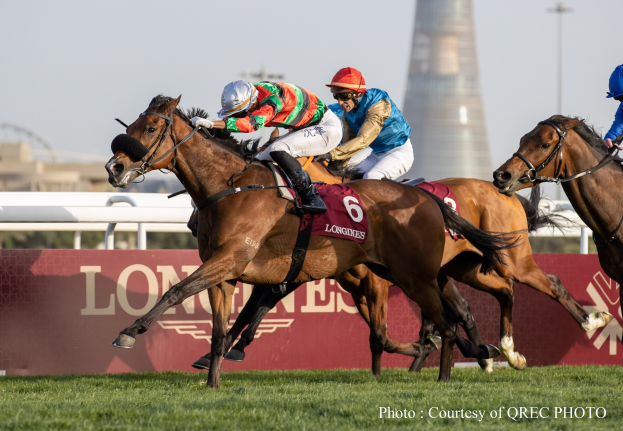 Drei Jockeys auf Pferden, die auf einer grasbedeckten Rennbahn mit einer Texttafel und einem Turm im Hintergrund unter einem klaren blauen Himmel galoppieren.
