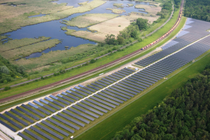 Luftaufnahme einer Solaranlage mit Solarpanelen auf einem Feld, umgeben von Bäumen, Gras, Pflanzen und Wasser, mit einem Zug auf einer nahen Eisenbahnbrücke.