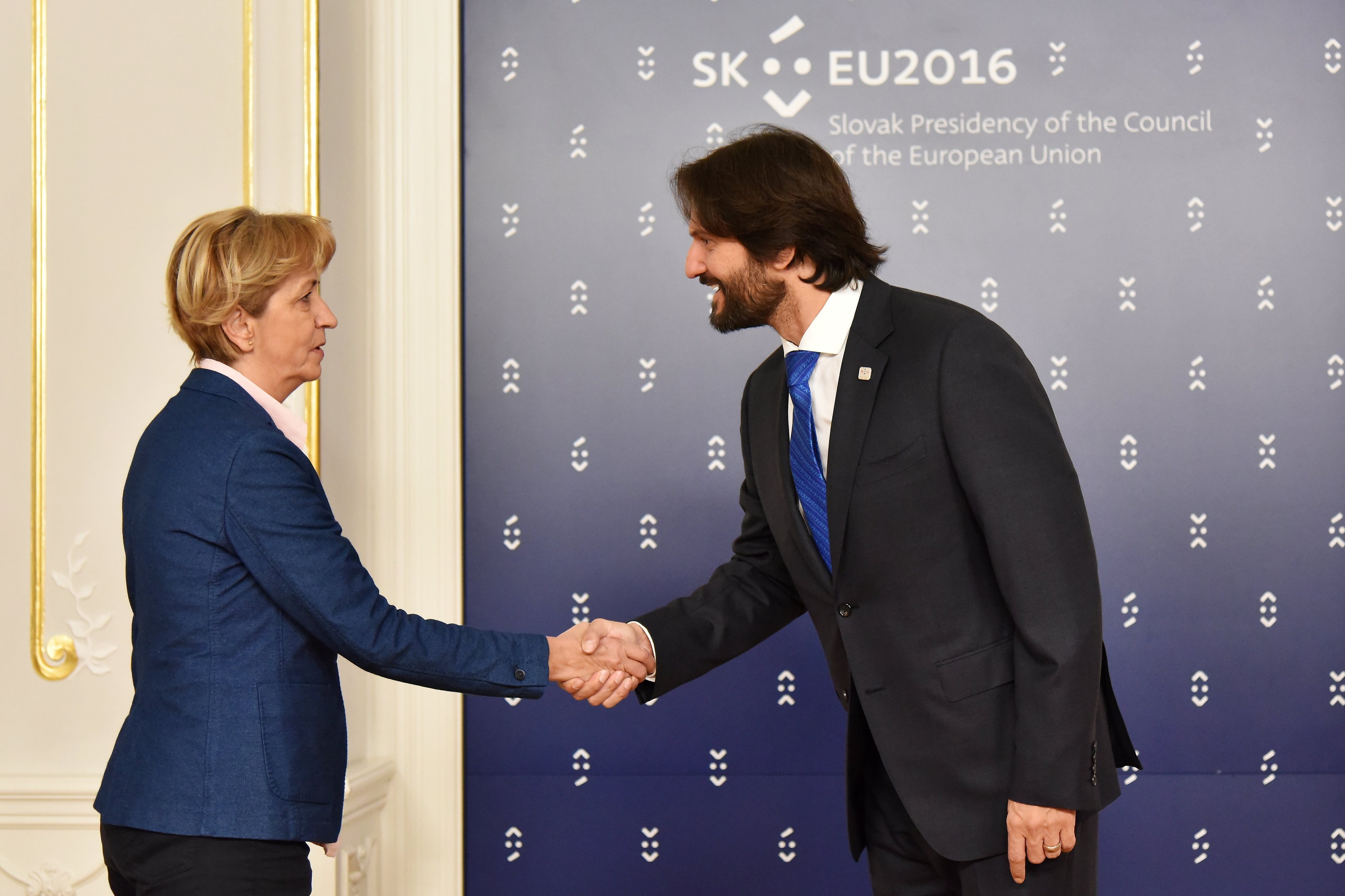 Bundeskanzlerin Angela Merkel und Bundespräsident Joachim Gauck schütteln sich die Hände vor einer Wand mit einem 'EU-Gipfel'-Schild und einer Tür im Hintergrund, beide lächeln.