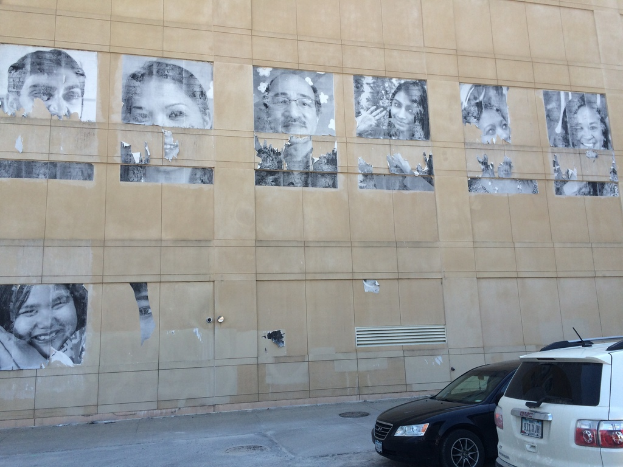 Ein Gebäude mit Autos auf der Straße davor, das Gullideckel und Plakate von Menschen an der Wand im Hintergrund zeigt.