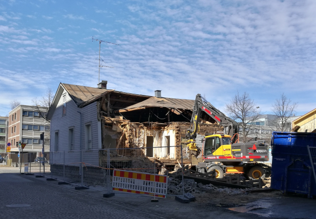 Baustelle mit einem großen Gebäude, das abgerissen wird, mit einem Kran, Zaun, Absperrung, Steinen, Container, Bäumen, Pfählen, Schild, Straßenpfosten, Fahrzeugen, Gebäuden mit Fenstern und einem bewölkten Himmel.