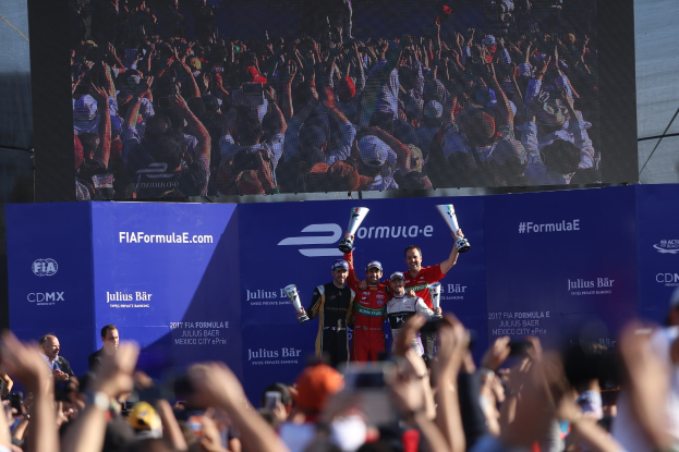 Podium mit einer Gruppe von Menschen, die Tassen halten, Hintergrund zeigt "FIA Formula E Mexico City" Branding, Bildschirm und Himmel.