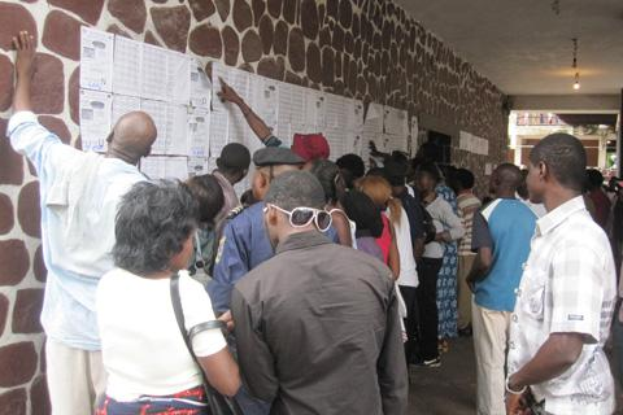 Eine Gruppe von Menschen steht vor einer Wand, die mit Papieren bedeckt ist, beleuchtet von Deckenlampen, wobei einige Personen Taschen tragen, was darauf hindeutet, dass sie wählen.