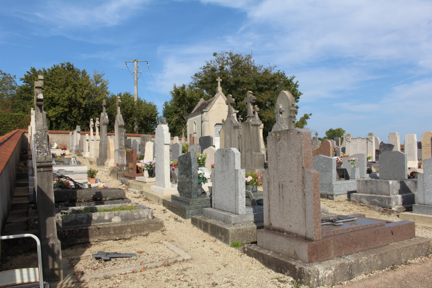 Ein Friedhof mit zahlreichen Grabsteinen und Blumen im Vordergrund, ein Gebäude im Hintergrund, Bäume, ein Strommast mit Drähten und ein bewölkter Himmel.